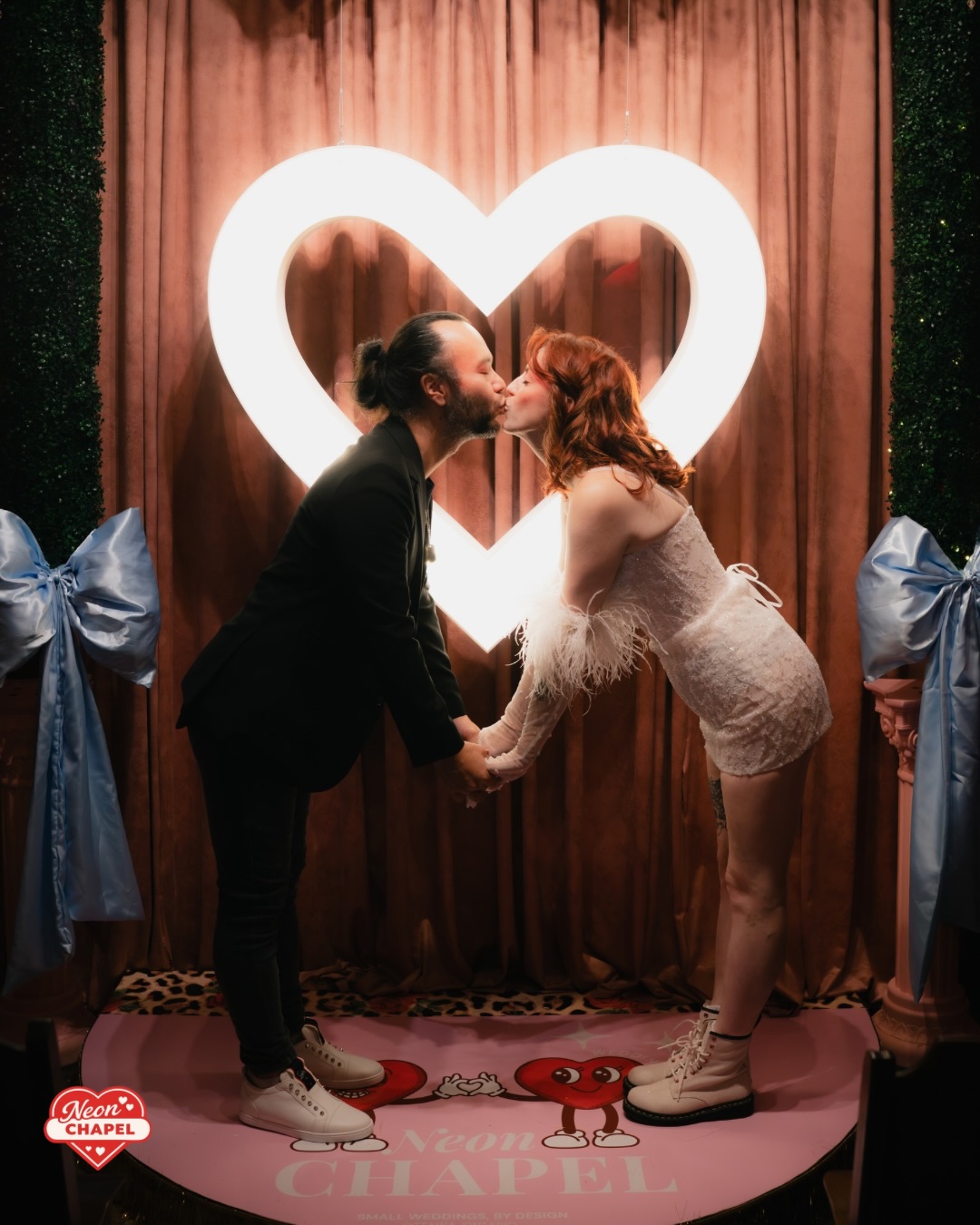 A couple kissing on the pink Neon Chapel stage beneath a glowing white heart, framed by pale blue satin bows