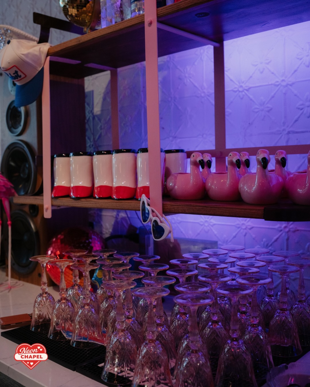 Bar shelf of pink flamingo cups and heart sunglasses above rows of upturned crystal glasses