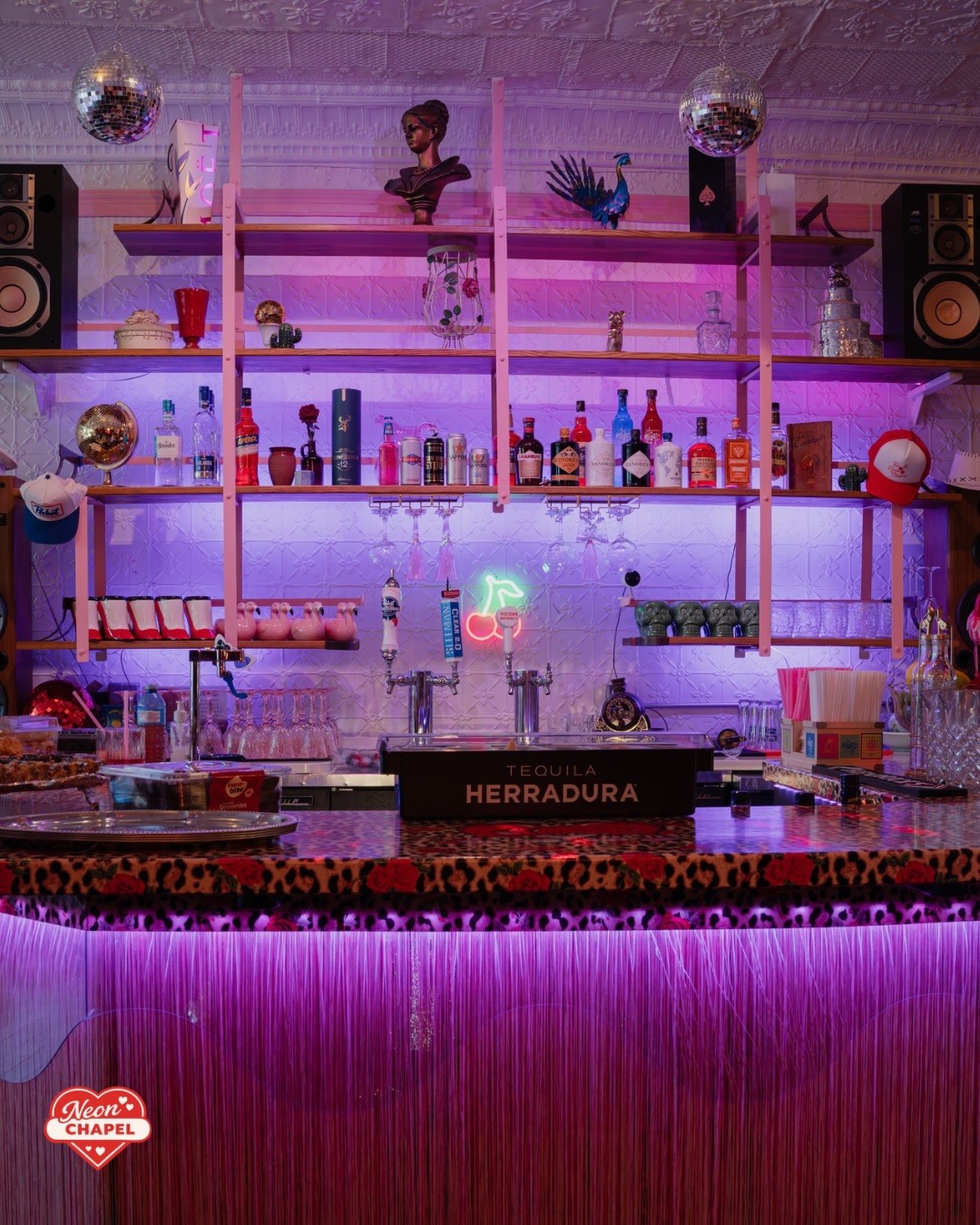 The bar glowing purple, with a neon cherry sign, disco balls, leopard-and-rose bartop and pink fringe