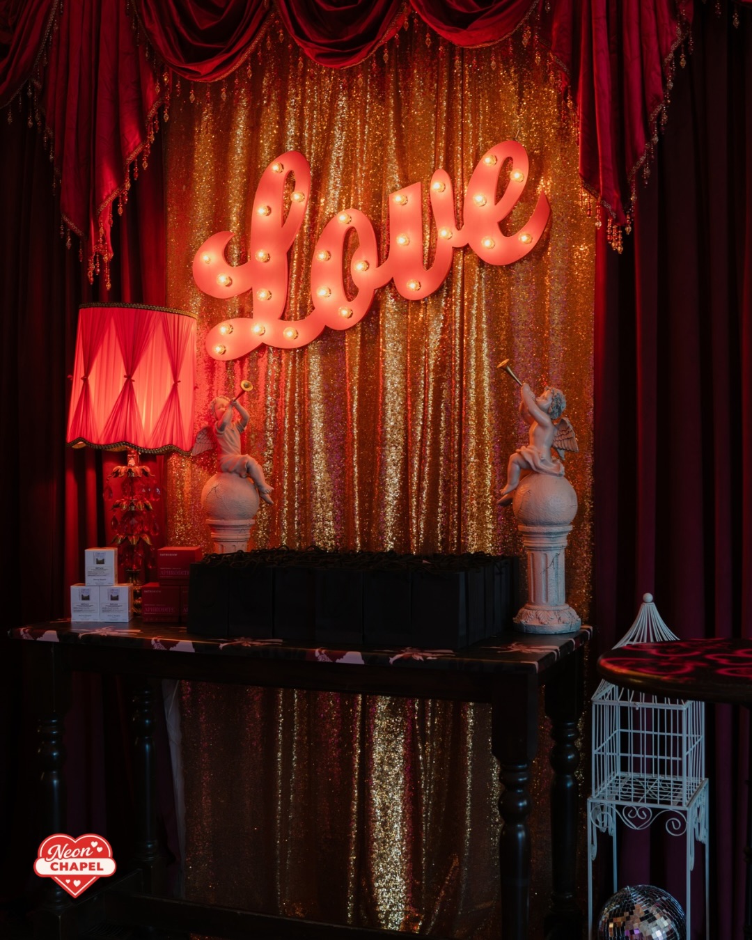 Light-up script Love sign over a gold sequin backdrop, flanked by cherub statues and a red fringed lamp